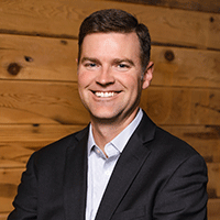 Headshot of a smiling man in a suit and against a wood wall background.