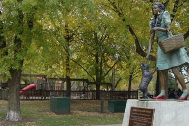 Bronze statue of Dorothy and Toto from the Wizard of Oz in a park. Playground in the background.
