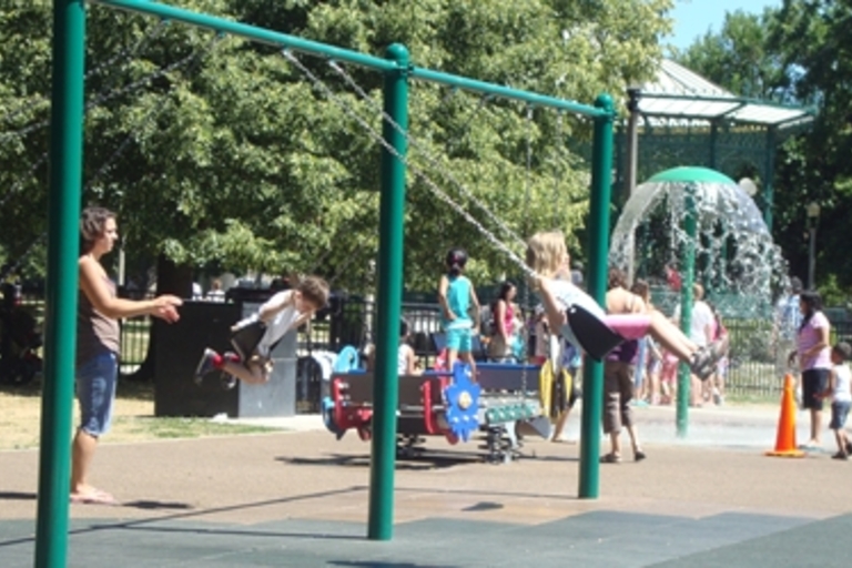 Children play on swings and at a splash pad on a sunny day.
