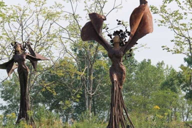 Two metal sculptures of winged female figures stand among trees.
