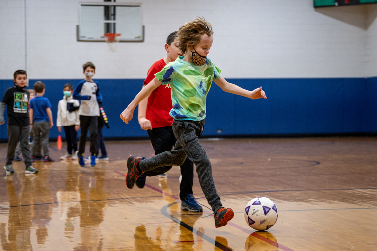Child wearing a mask kicks a soccer ball in a gym. Other children in the background.