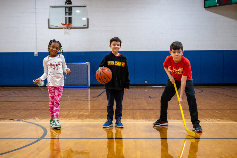 Three children with jump rope, basketball, and hockey stick in a gym.