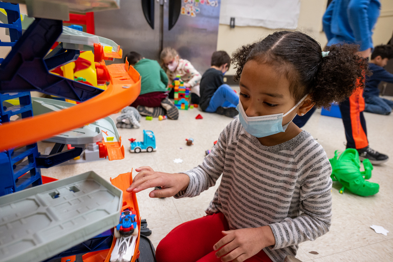 A masked child plays with a toy car and ramp. Other children play in the background.