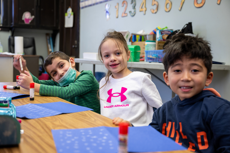 Three young students smile at the camera in a classroom.