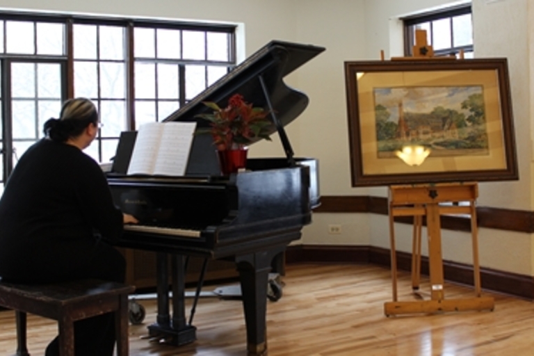 Pianist plays grand piano next to painting on easel.
