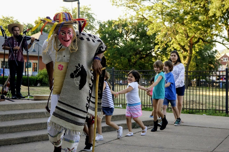 A masked traditional dancer and children dance in a park as musicians play in the background.
