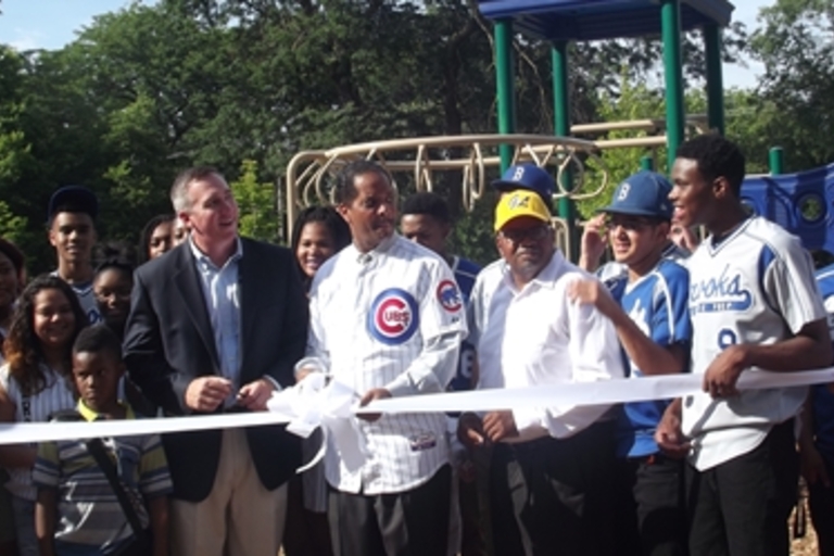 Ribbon-cutting ceremony at a playground with community members and baseball players.
