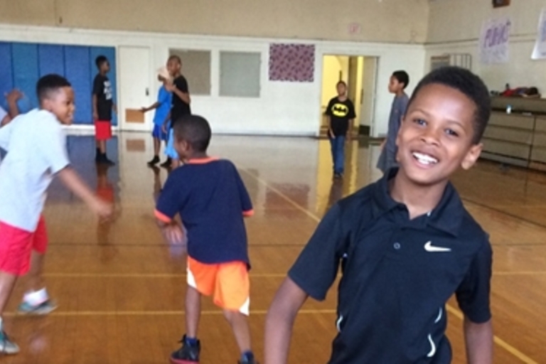 Smiling boy in black shirt on basketball court with other children.
