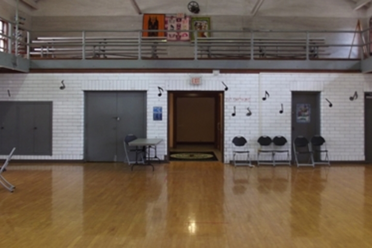 Empty dance studio with polished wood floor, white tile walls, and musical note decorations. A small mezzanine level is visible.