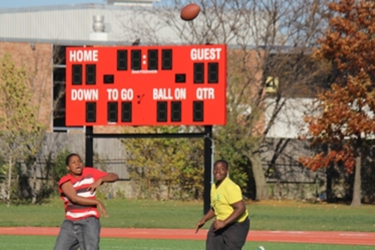 Two people play catch with a football on a field near a scoreboard.