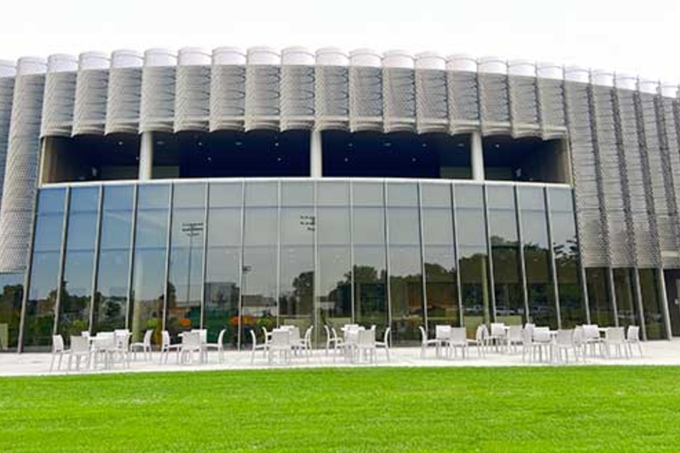 Modern building with glass facade and unique patterned metal exterior, patio seating in front, and green lawn.