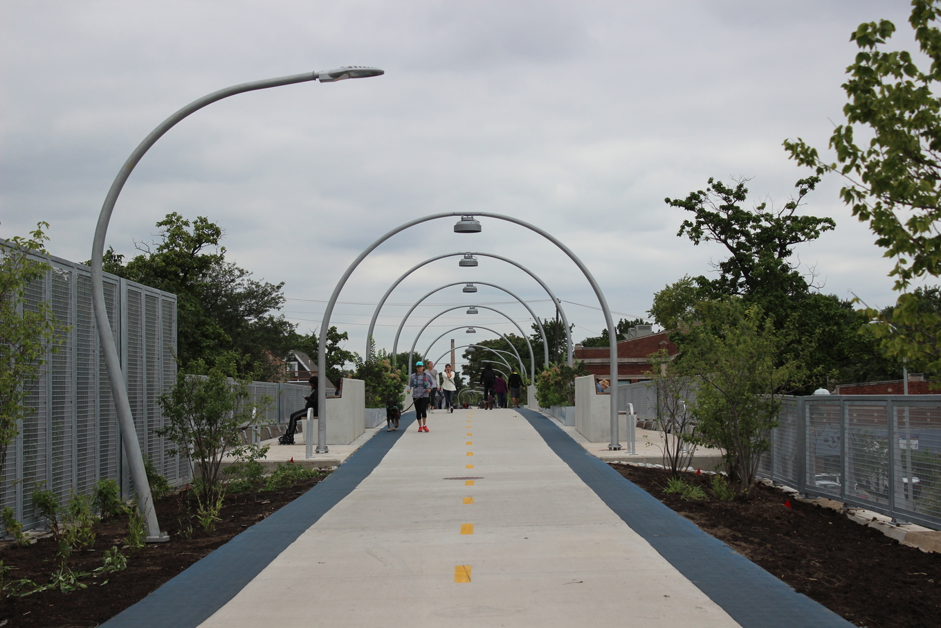 A paved path with a series of arched lampposts stretches into the distance, with a few people walking and cycling.