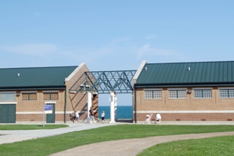 Two brick buildings with green roofs connected by a metal trellis archway. People walk near the buildings and through the archway towards the lake.