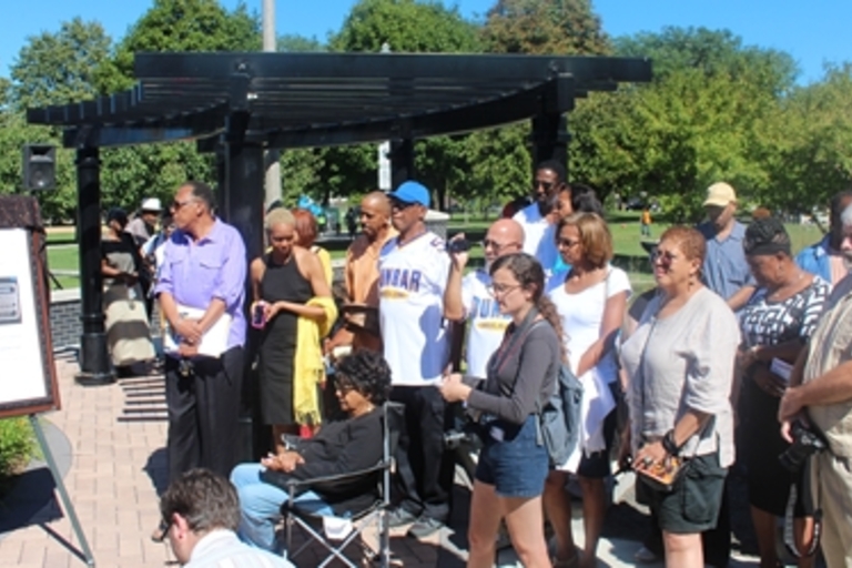 Group gathers at outdoor ceremony with a historical information board.