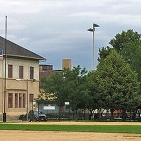 Tan school building with US flag at half-mast and baseball field.
