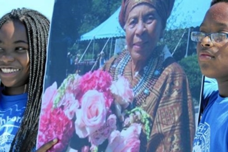 Two children hold a large photo of an older woman holding a bouquet of pink roses.
