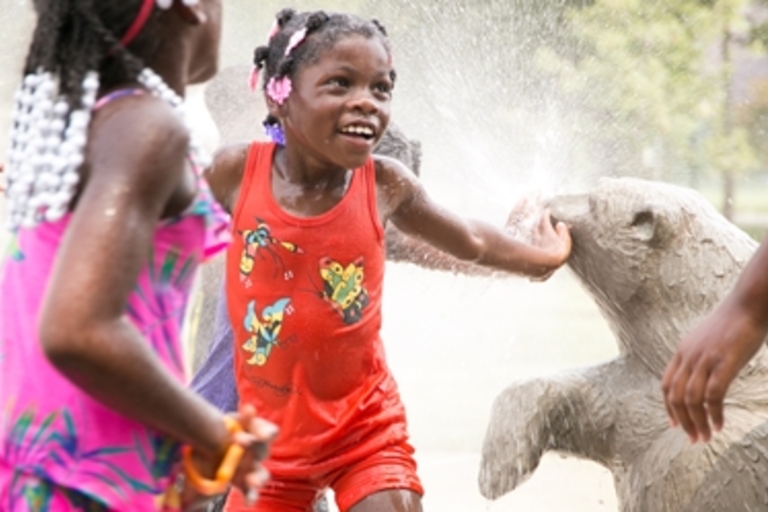 Children play in a splash park with a bear sculpture.
