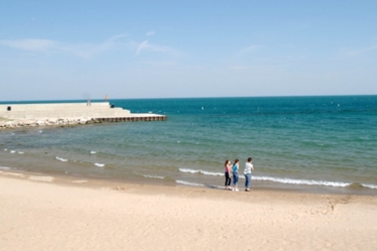 Three people stand on a sandy beach by the calm, blue water. A breakwater juts out in the background.