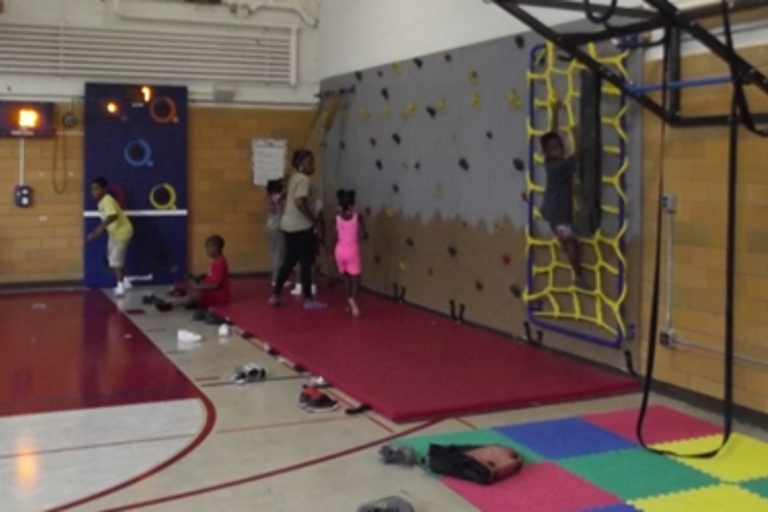 Children play in a school gymnasium. Some climb a rope net while others play with targets on the opposite wall.