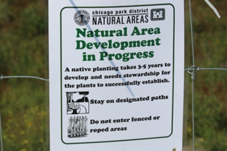 Sign on a wire fence reads "Natural Area Development in Progress".