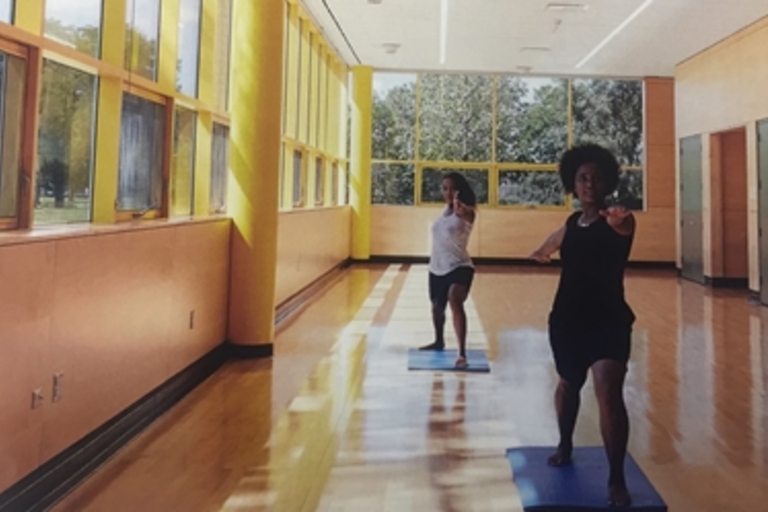 Two people practice yoga in a brightly lit room with large windows.
