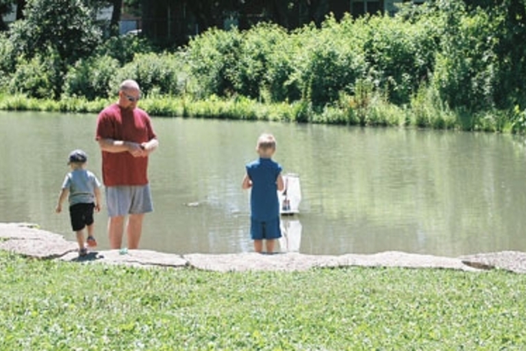 Dad and two kids playing with toy sailboat on pond.
