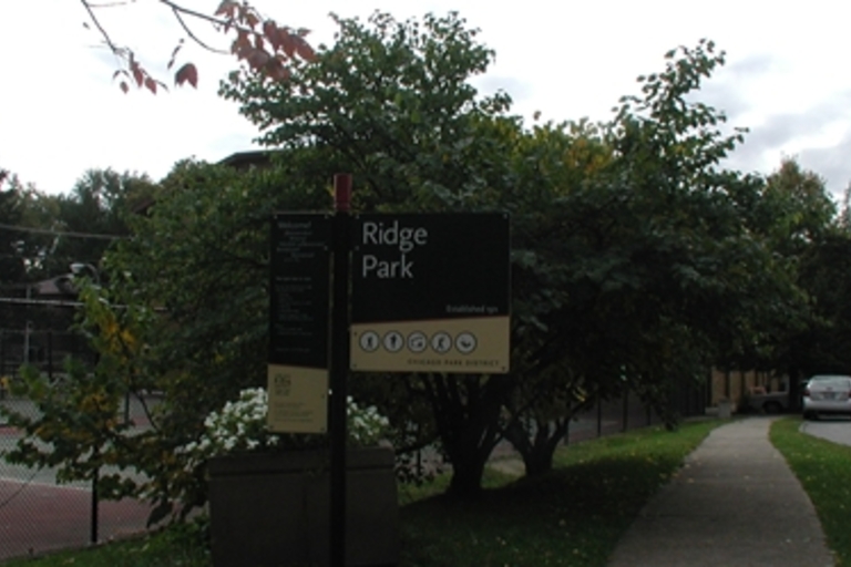 Ridge Park sign with a tennis court, trees, and a building in the background.