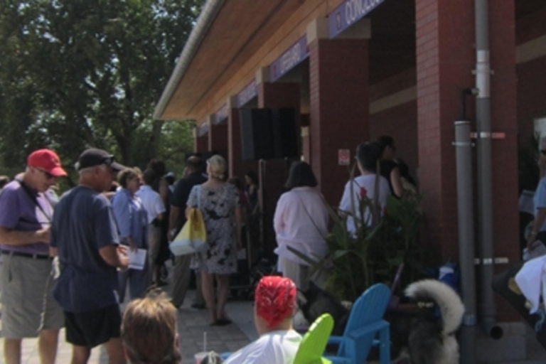Crowd of people waiting in line outside a brick building on a sunny day.
