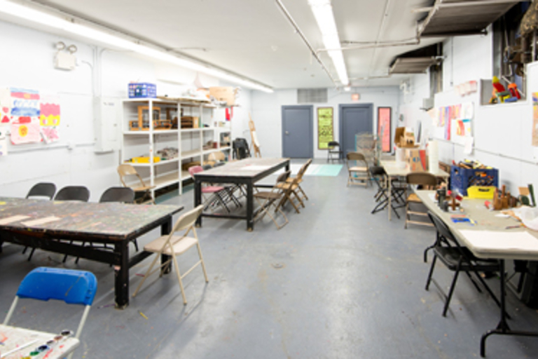 A brightly lit art classroom with several tables, chairs, and children's artwork displayed.