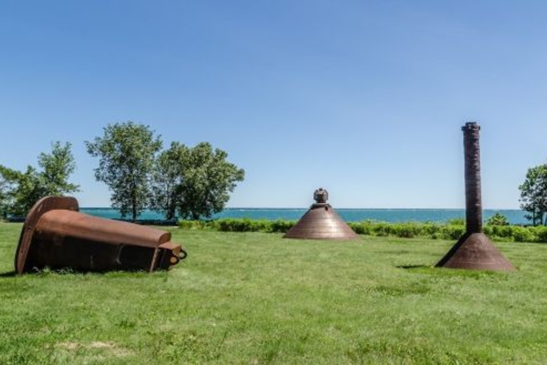 Rusty metal sculptures in a grassy field overlooking water.
