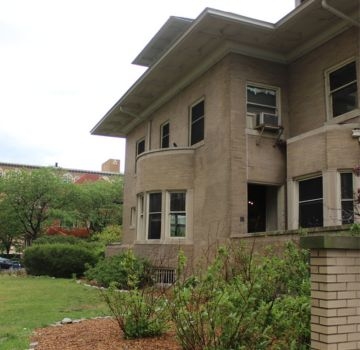 Tan brick prairie school house with rounded porch and shrubs.