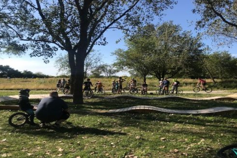 Cyclists of all ages gather at a bike park with rolling wooden ramps.
