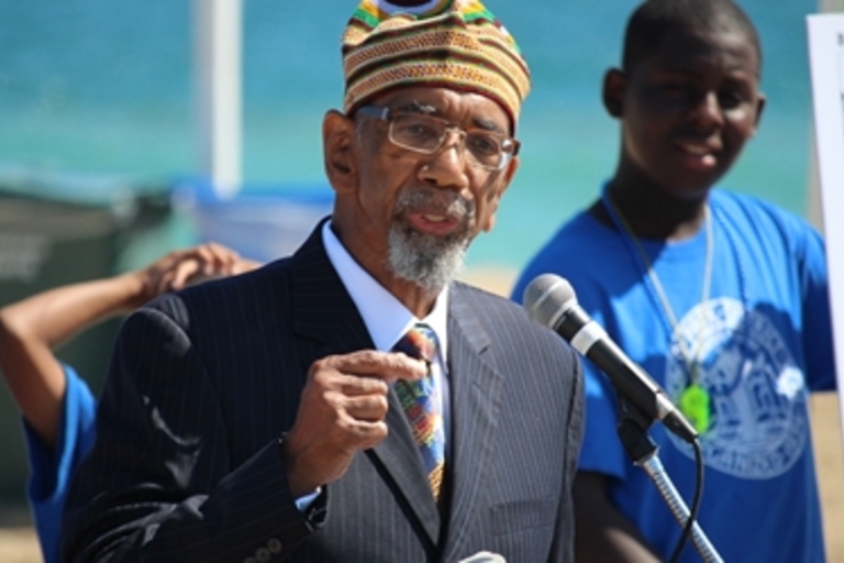 Man in suit and colorful kufi speaking at a microphone.