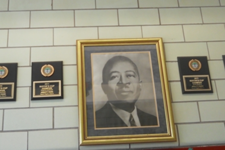 Framed portrait of a man and four Mayor's Cup Champion plaques on a tiled wall.
