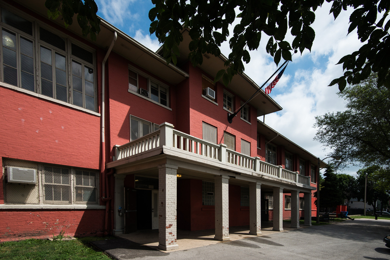 Red brick building with a covered entryway supported by white pillars and a small balcony. An American flag hangs from the roof.