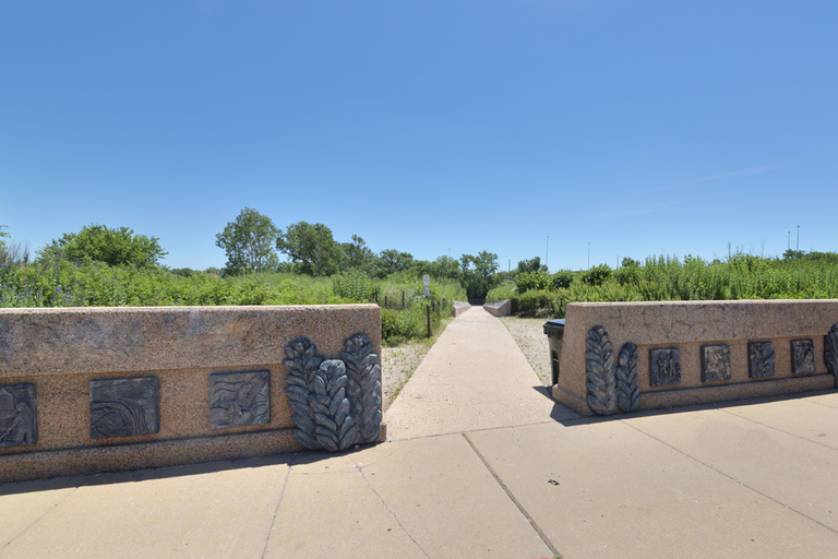 A paved path leads through a nature preserve, passing between low concrete walls decorated with nature-themed reliefs.