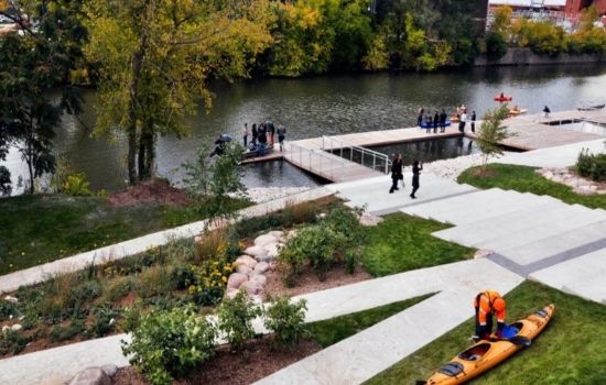 Riverfront park with docks, walkways, and people kayaking.