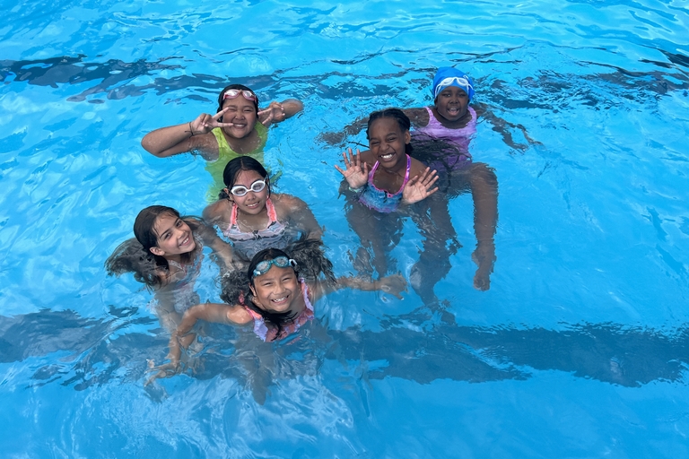 Six children smile and play in a bright blue swimming pool. Most wear goggles.