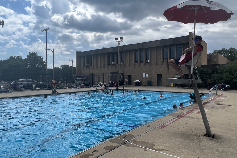 Outdoor community pool with people swimming on a partly cloudy day. A lifeguard sits in a chair under an umbrella.