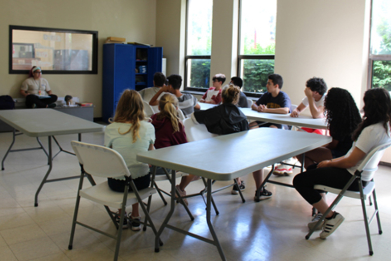 Students sit at tables and listen to an instructor.