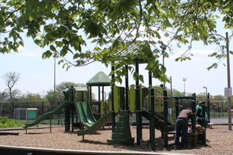 Green playground structure on wood chips with adults nearby.
