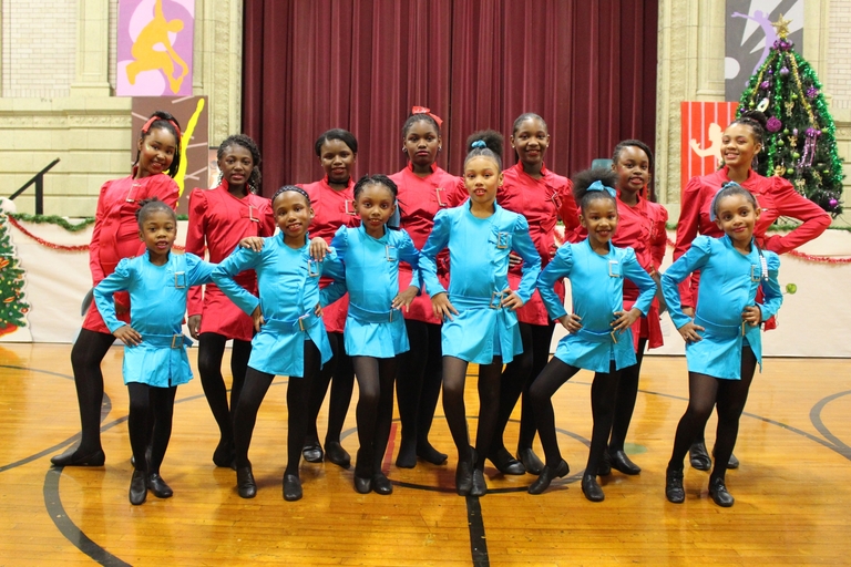A dance troupe of young girls in red and teal costumes pose on a gymnasium floor. A decorated Christmas tree stands in the background.