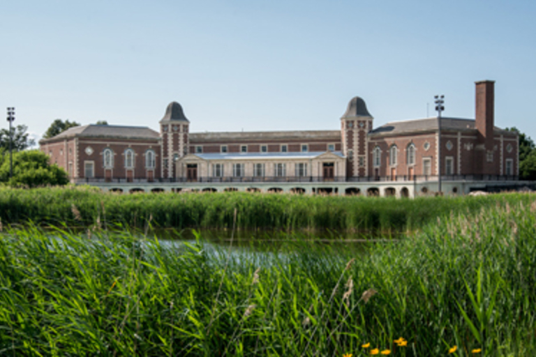 Brick building with tall arched windows viewed across a pond with tall grass.