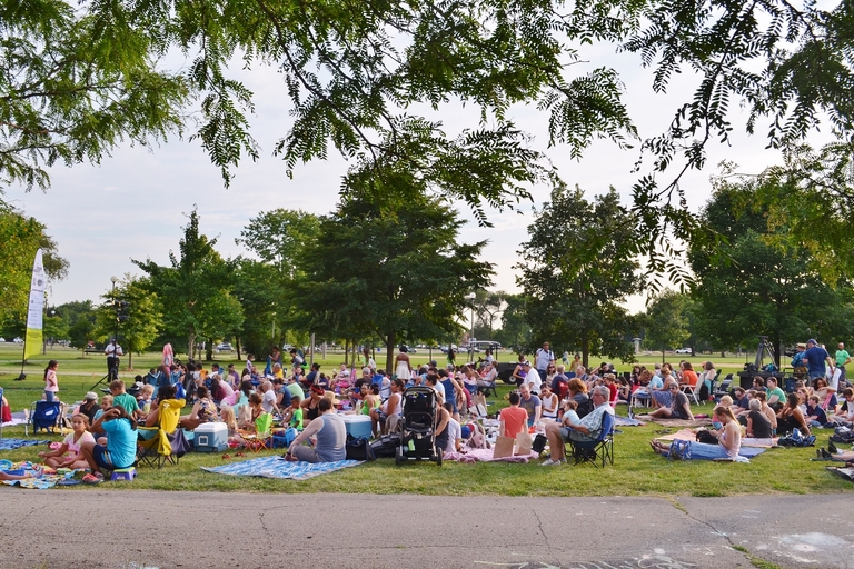 A crowd of people sit on blankets and lawn chairs in a park, enjoying an outdoor event.  Trees frame the scene.