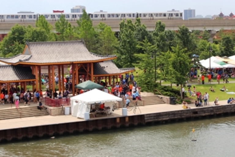 Crowds gather at a Chinese pavilion and along a riverwalk during a festival. A train passes on an elevated track.