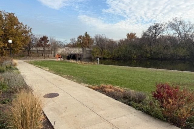 A concrete walkway beside a grassy bank, river, and bridge under a partly cloudy sky.