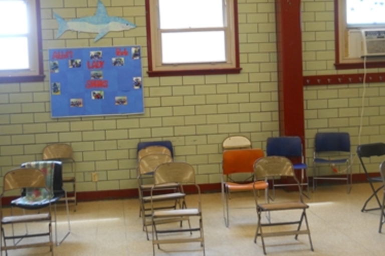 Empty metal chairs in a classroom with pale green tile walls.