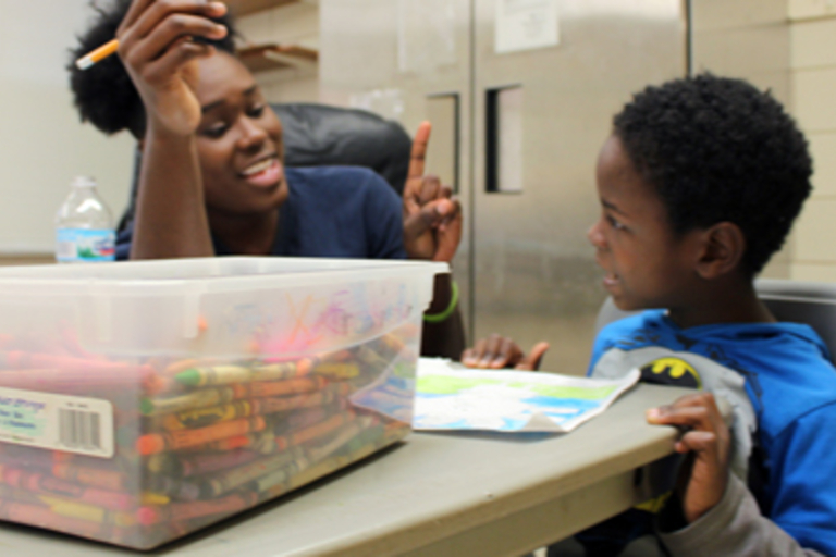 Teacher and student look at drawing, crayons in container between them.