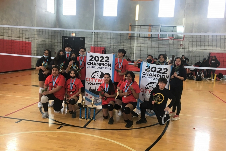 A youth volleyball team poses with their championship banner and trophies.
