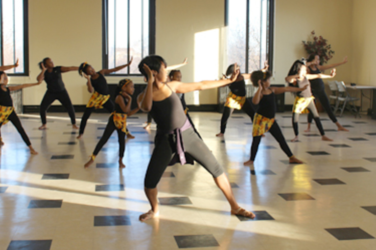 Dancers in black and gold rehearse a routine in a sunlit studio.

